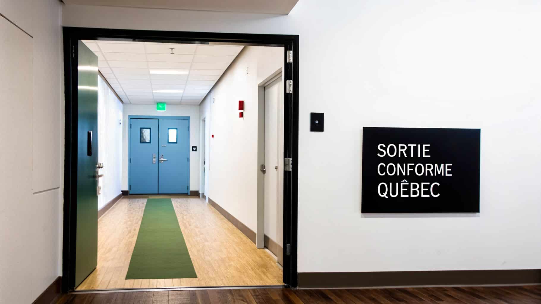 Un couloir blanc avec une porte verte ouverte sur la gauche, menant à des portes bleues au fond. Un panneau noir sur le mur de droite indique "SORTIE CONFORME QUÉBEC".